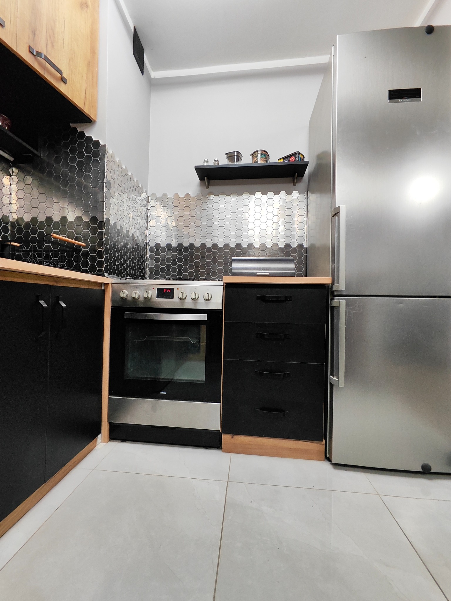 Nowoczesna kuchnia z czarnymi szafkami z drewnianymi akcentami, srebrną lodówką i piekarnikiem Amica, oraz heksagonalnymi płytkami na ścianie.