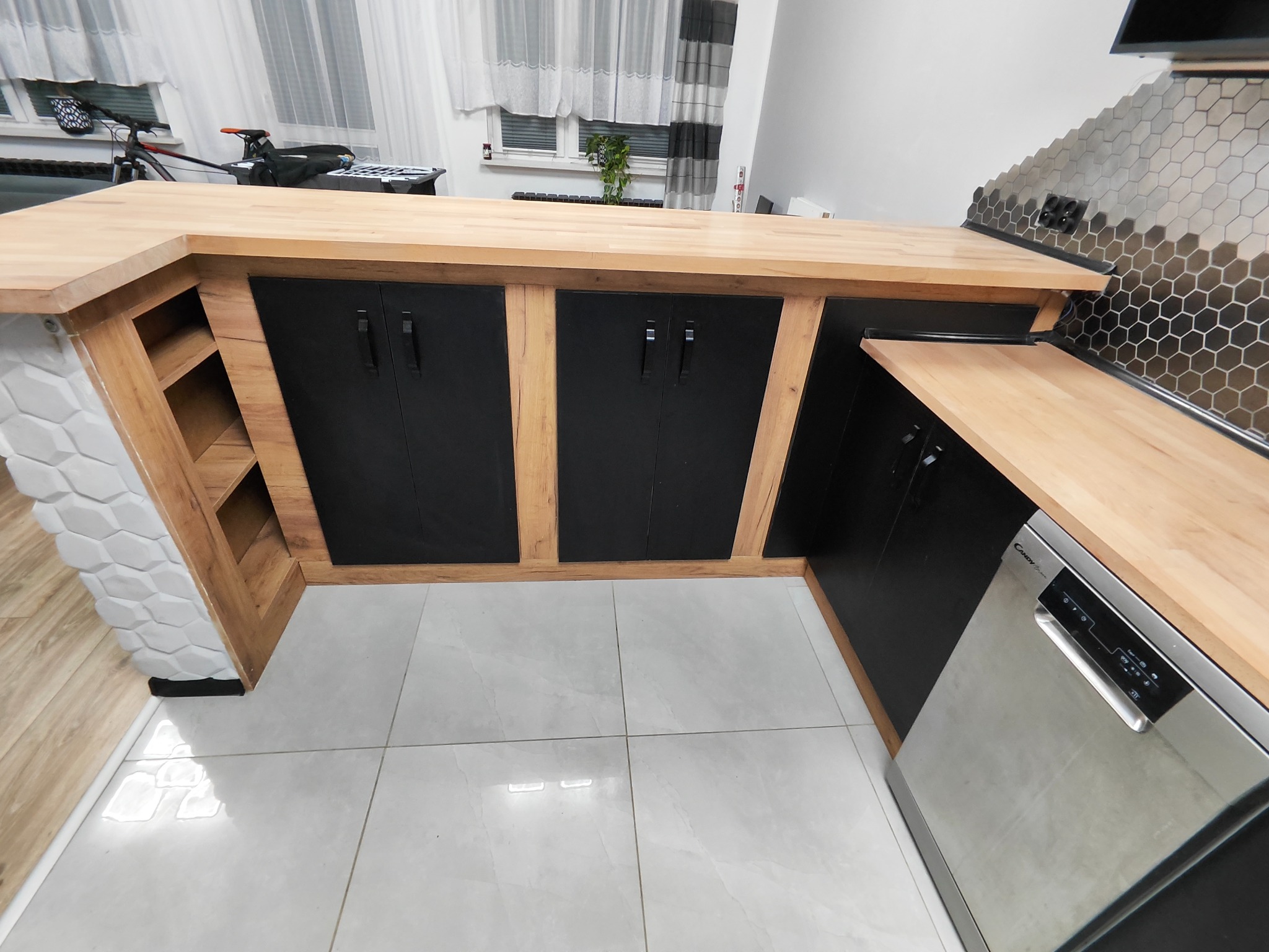 Nowoczesna szafa na wymiar z drewnianym blatem i czarnymi frontami, wkomponowana w aneks kuchenny z szarymi płytkami heksagonalnymi na ścianie i białą mozaiką na boku szafy.