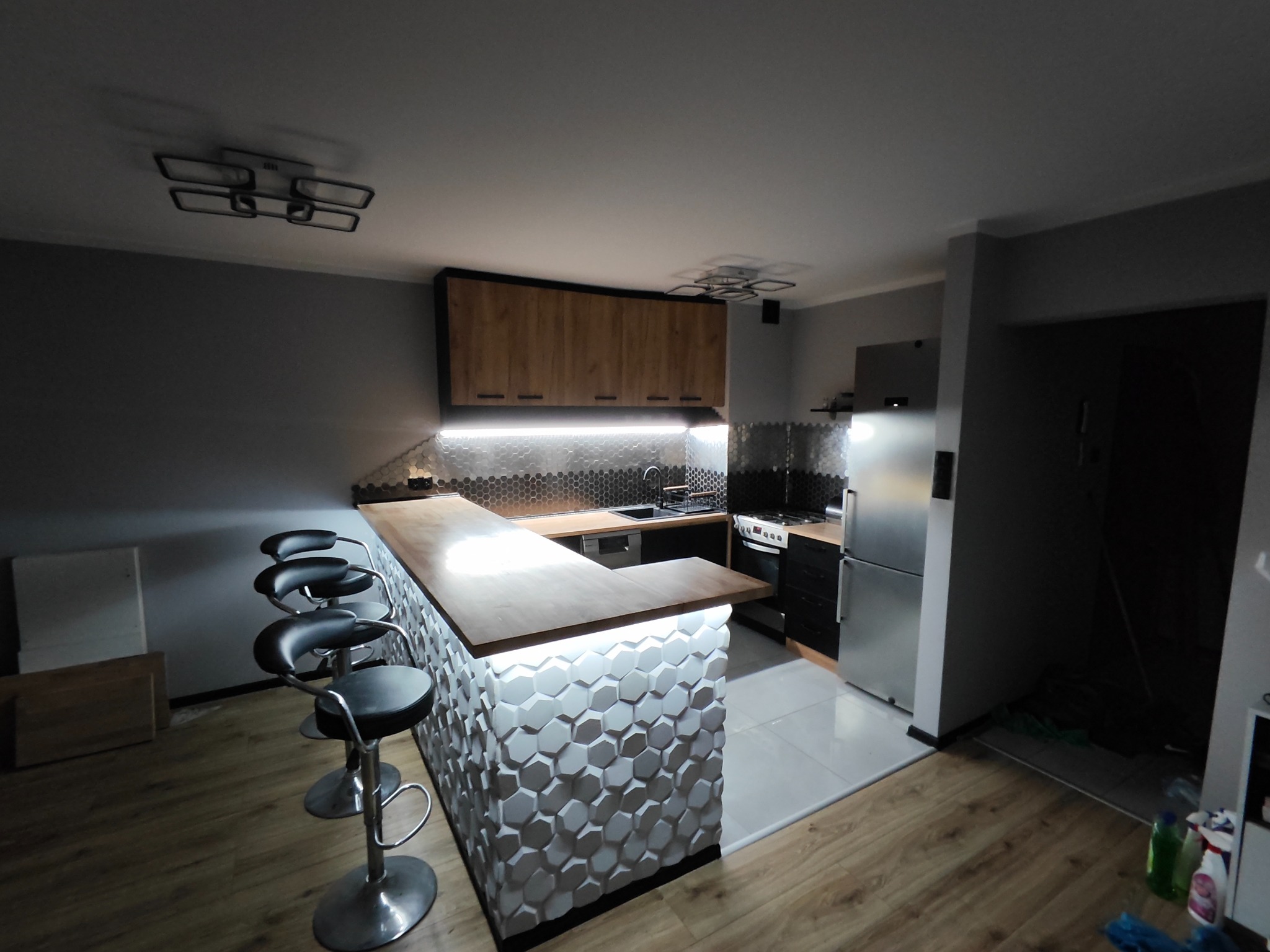 Nowoczesna kuchnia z wyspą wykończoną trójwymiarowymi panelami, oświetleniem LED pod blatem i czterema hokerami.