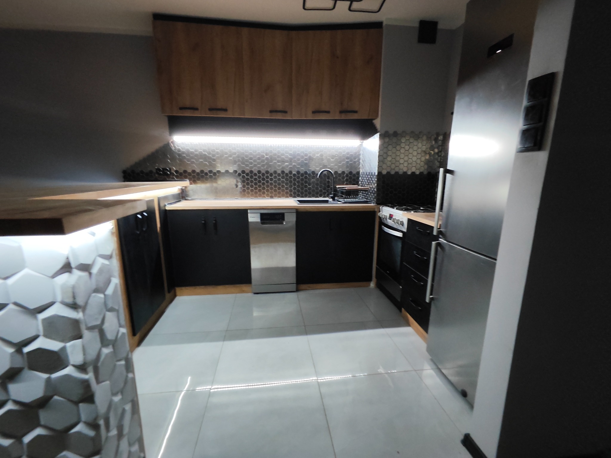 Nowoczesna kuchnia z drewnianymi szafkami wiszącymi, czarnymi szafkami dolnymi, srebrnymi płytkami heksagonalnymi nad blatem i białymi płytkami podłogowymi.