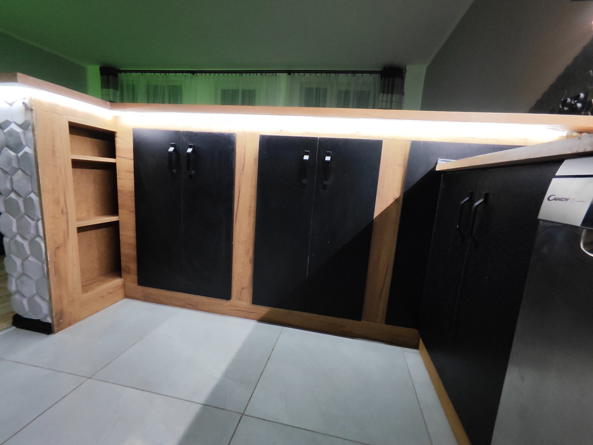 Nowoczesna zabudowa kuchenna z czarnymi frontami i drewnianymi ramami, podświetlana listwą LED, uzupełniona dekoracyjnym panelem ściennym o strukturze plastra miodu.