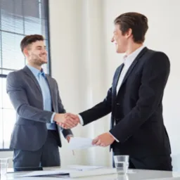 Dwóch uśmiechniętych mężczyzn w garniturach w trakcie uścisku dłoni nad dokumentami na biurku w jasnym biurze.