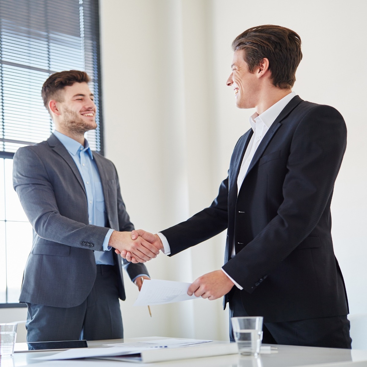 Dwóch uśmiechniętych mężczyzn w garniturach w trakcie uścisku dłoni nad dokumentami na biurku w jasnym biurze.