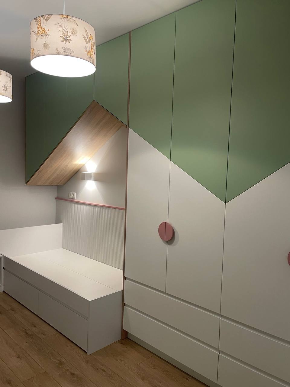 Zabudowa meblowa w pokoju dziecięcym z szafą w kolorach zieleni i bieli, zintegrowana z łóżkiem i oświetleniem punktowym, wykończona elementami w kolorze różowym.
