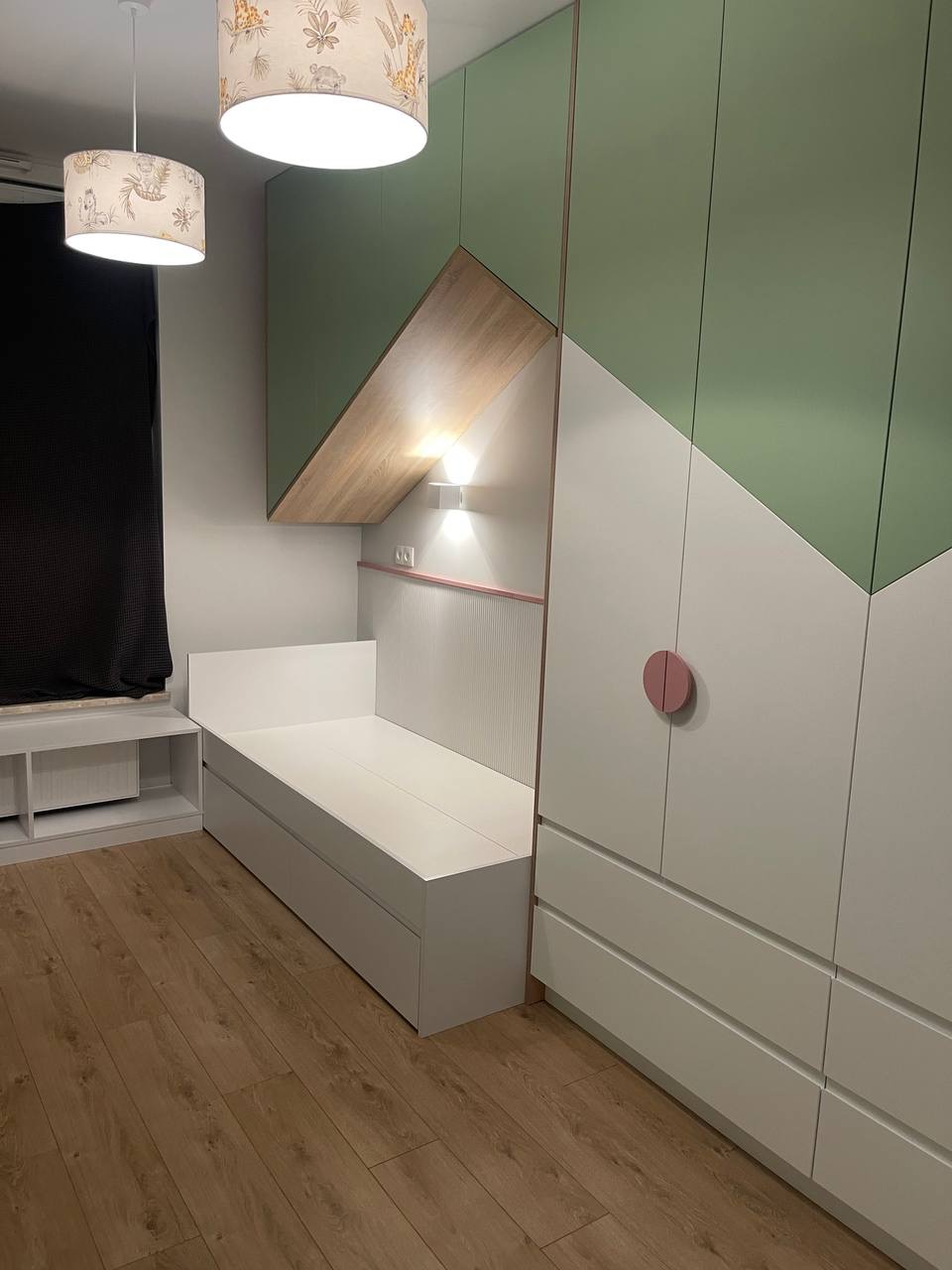 Pokój dziecięcy z zabudową meblową: białe łóżko z szufladami, szafa w odcieniach bieli i zieleni z okrągłym, różowym uchwytem, nad łóżkiem półka z oświetleniem, drewniana podłoga, lampy z motywem...