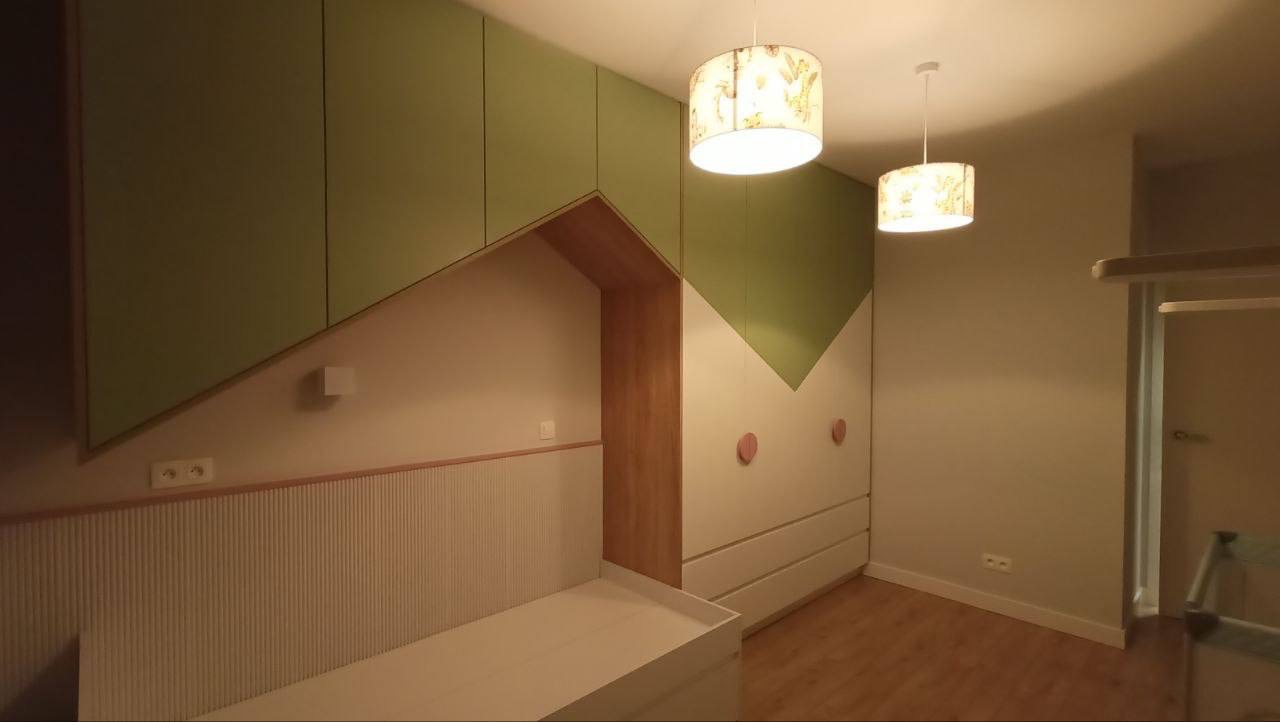 Pokój dziecięcy z zabudową meblową: szafa z zielonymi akcentami i drewnianą wnęką nad łóżkiem, uzupełniony o lampy z motywami roślinnymi.