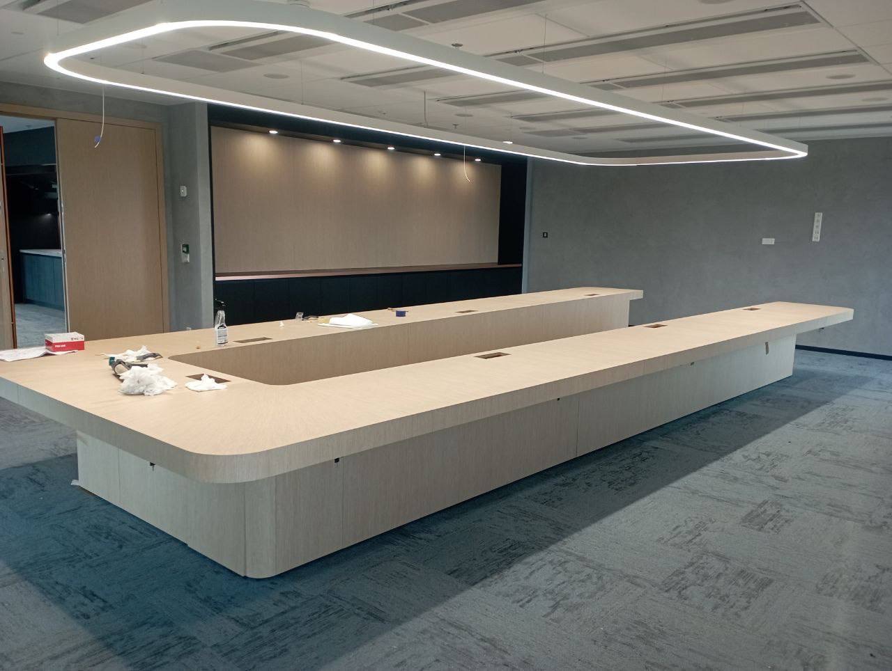 Długa, jasna, drewniana sala konferencyjna w biurze, z widocznymi elementami montażu, takimi jak butelka wody i ściereczki, oświetlona nowoczesną lampą sufitową LED.