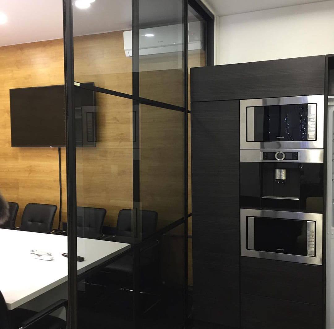 Nowoczesna kuchnia w biurze z zabudową w ciemnym drewnie, zawierająca piekarnik, ekspres do kawy i mikrofalówkę, oddzielona szklaną ścianką od sali konferencyjnej z białym stołem i czarnymi krzesłami.