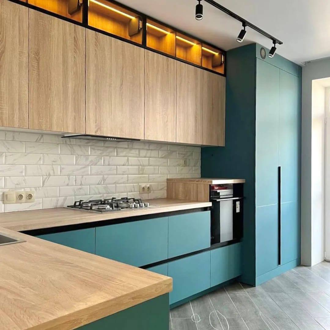 Nowoczesna kuchnia z drewnianymi szafkami górnymi z podświetleniem LED, turkusowymi szafkami dolnymi i białymi płytkami imitującymi marmur na ścianie, oświetlenie szynowe na suficie.