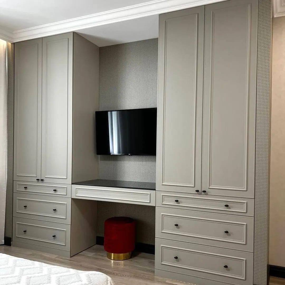 Szara zabudowa meblowa z szafkami, szufladami i biurkiem, wnęka z telewizorem i tapetą w drobną kratkę, czerwony stołek z metalową podstawą.