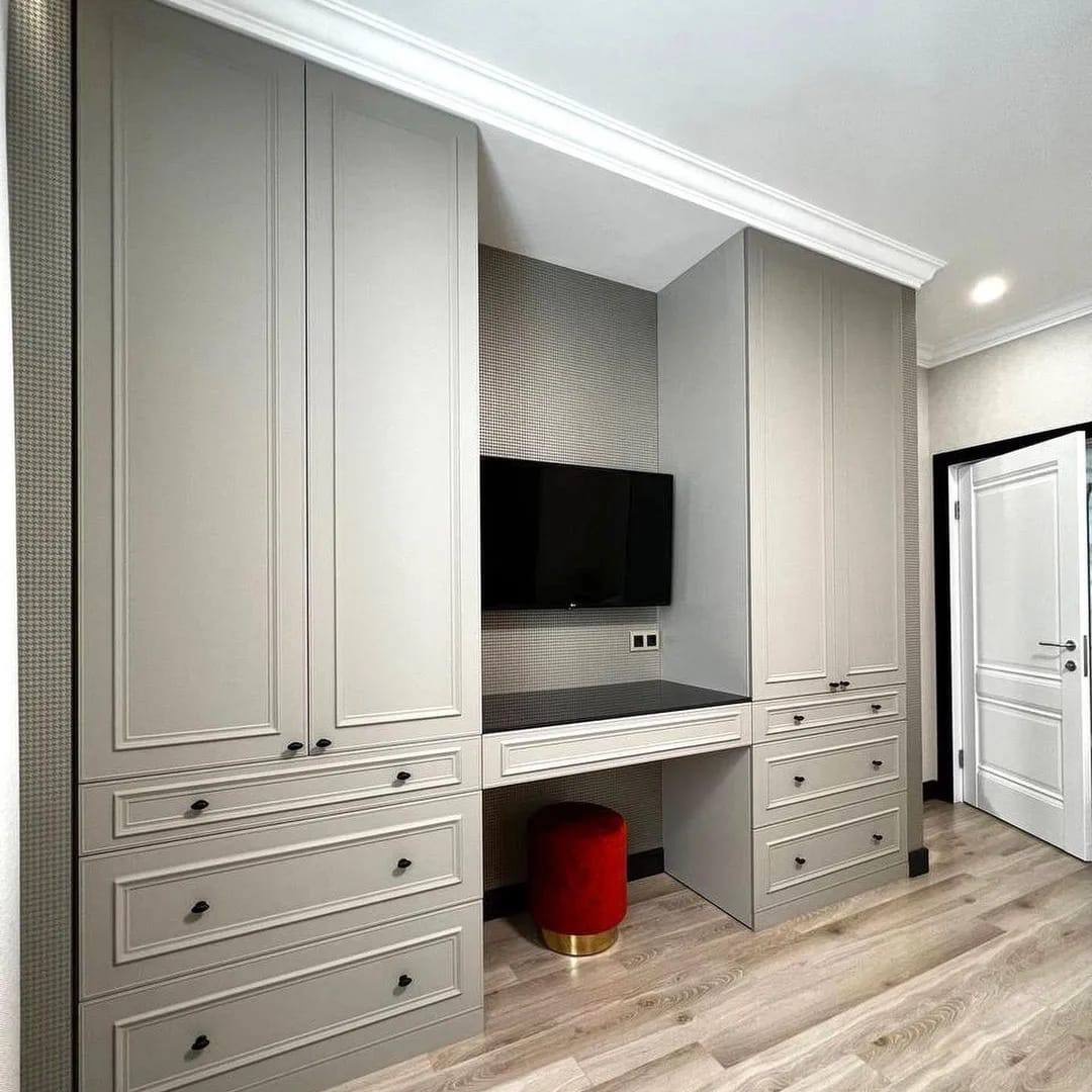 Szara zabudowa meblowa na wymiar z biurkiem i miejscem na telewizor, czerwony welurowy taboret ze złotą podstawą, jasna podłoga, dekoracyjne listwy przysufitowe.