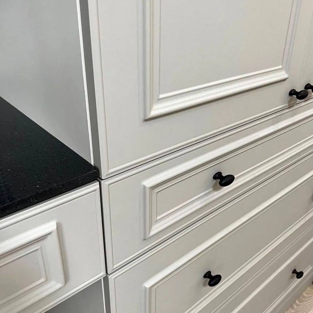 Szczegółowe ujęcie białej zabudowy kuchennej z frezowanymi frontami szuflad i szafek, czarny blat z drobnymi, błyszczącymi inkluzjami, oraz matowe, czarne gałki.