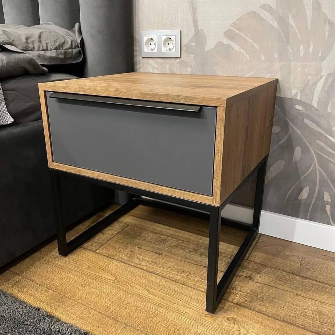 Stolik nocny z szarą szufladą i drewnianym korpusem na czarnych, metalowych nogach, widoczny na tle podłogi z paneli i ściany z motywem liści.