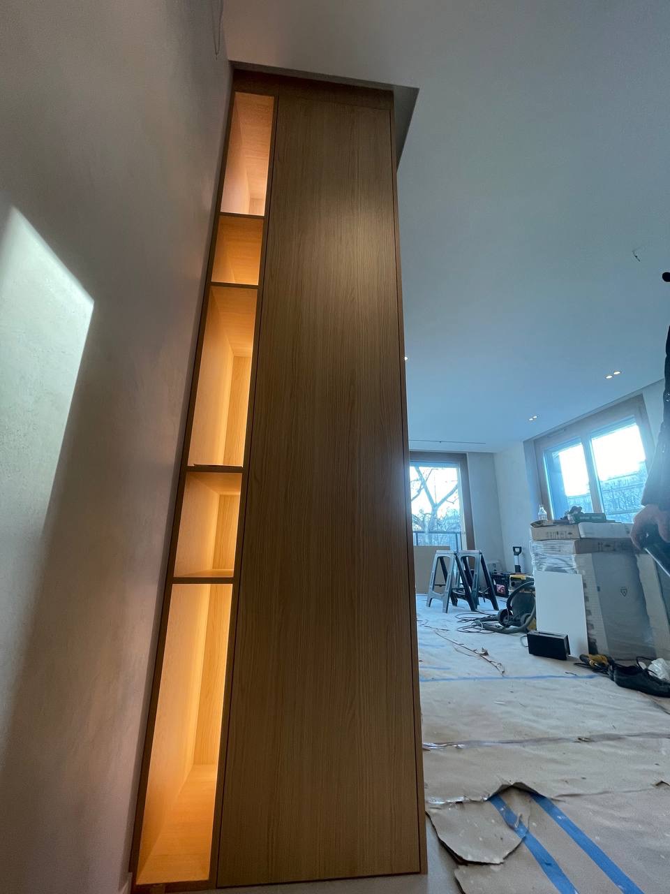 Wysoka, drewniana zabudowa meblowa z oświetlonymi półkami wnękowymi, w trakcie montażu, widoczne zabezpieczenia podłogi i narzędzia w tle.