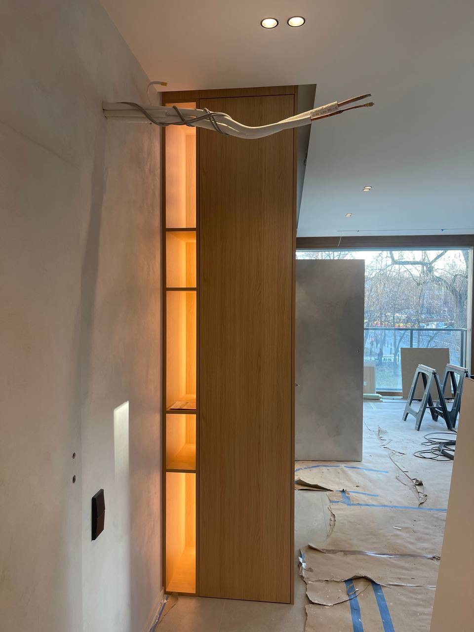 Wnęka z półkami podświetlona ciepłym światłem, z widocznymi przewodami instalacji klimatyzacji na górze, w trakcie prac wykończeniowych.