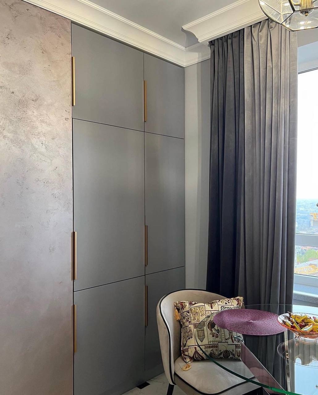 Nowoczesna kuchnia z zabudową szaf sięgającą sufitu, szare fronty z długimi, złotymi uchwytami, ściana wykończona tynkiem dekoracyjnym, obok okna zasłonięte grubymi, grafitowymi zasłonami, krzesło...