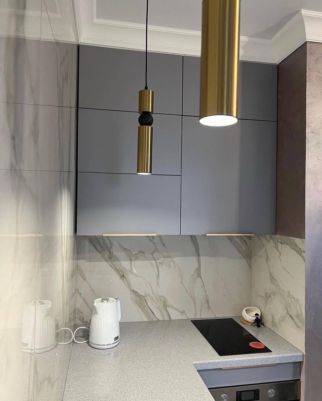 Szare szafki kuchenne z marmurowym backsplashiem, oświetlone złotymi lampami wiszącymi, z czajnikami na blacie i płytą indukcyjną.