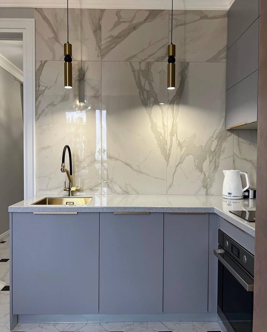 Nowoczesna kuchnia z szarymi szafkami, blatem z jasnego kamienia, marmurową ścianą i złotą armaturą oraz lampami wiszącymi.
