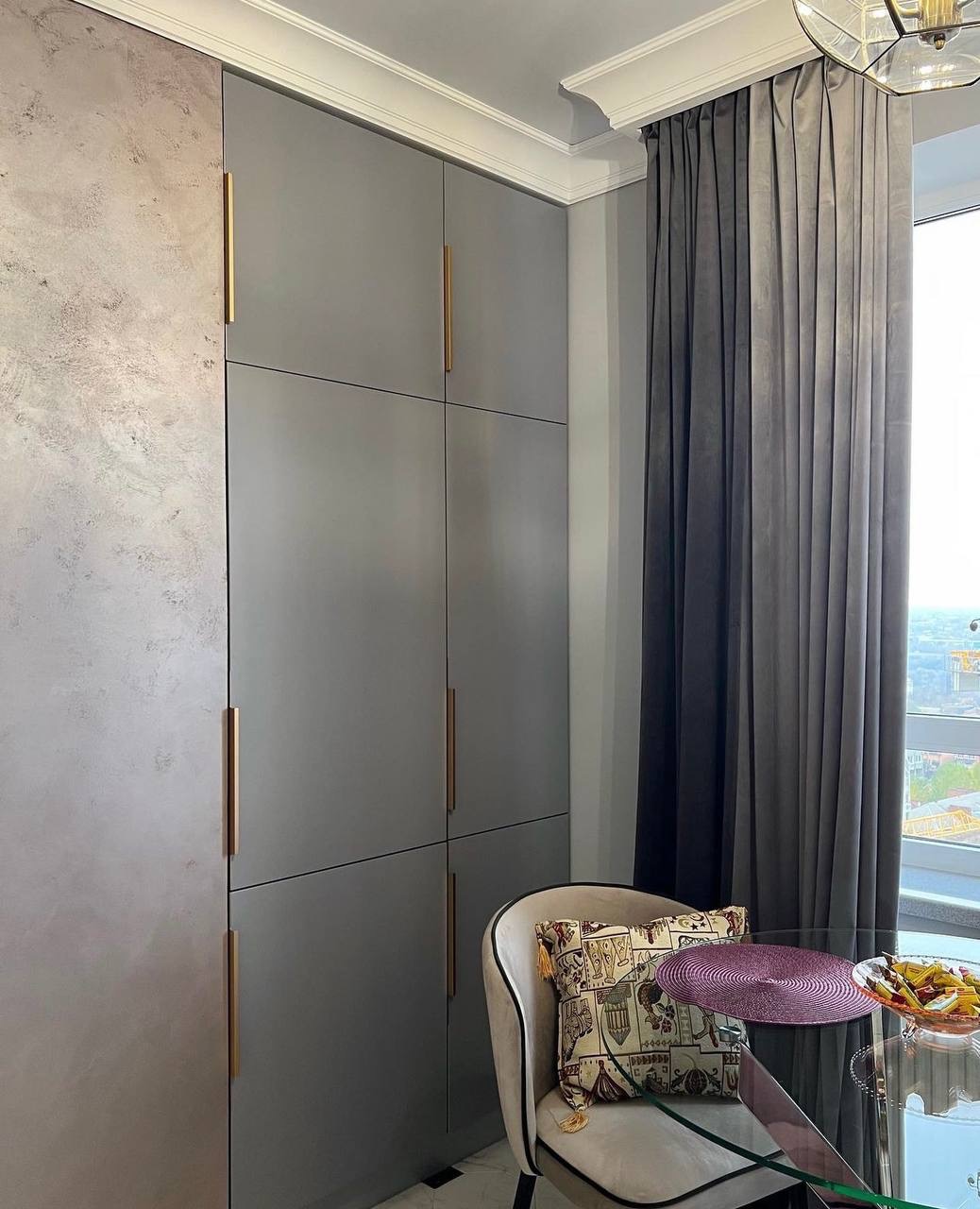 Szara zabudowa kuchenna z długimi, złotymi uchwytami, widoczny fragment szklanego stołu z krzesłem i dekoracyjną poduszką, zasłony w odcieniach szarości i widok z okna.