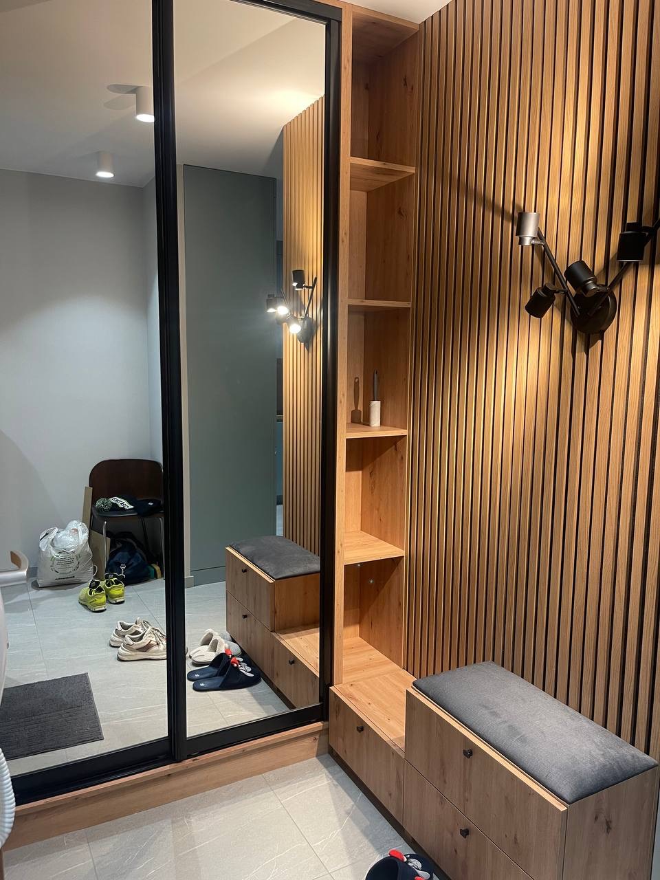 Nowoczesna zabudowa przedpokoju z lustrem w czarnej ramie, drewnianymi panelami ściennymi i szafką z siedziskiem w szarym kolorze.