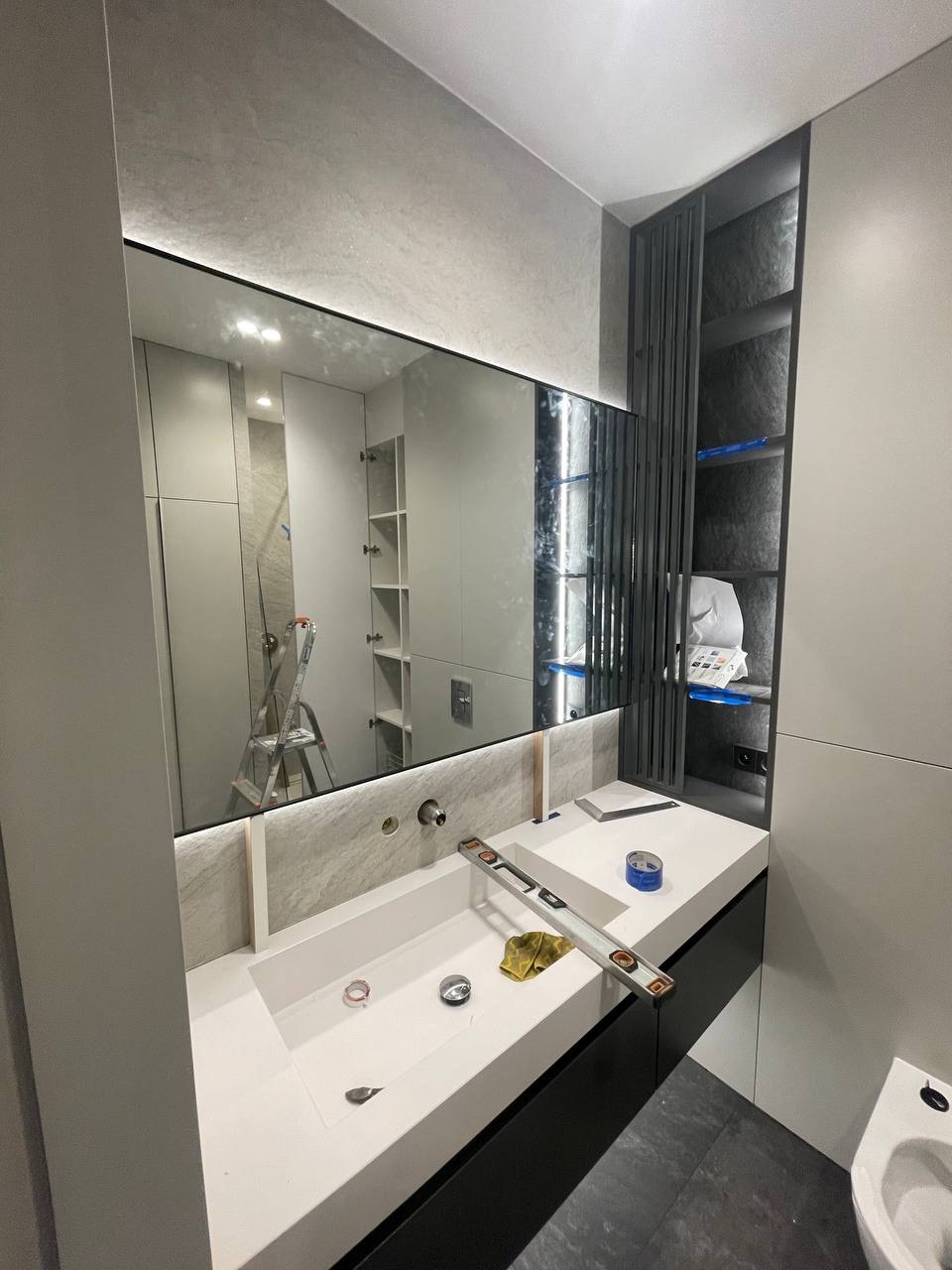 Nowoczesna łazienka w trakcie remontu: biała umywalka wpuszczona w blat, poziomica, lustro z podświetleniem LED odbijające drabinę, szara zabudowa meblowa z półkami.