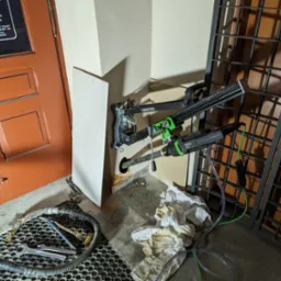 Wiertnica Eibenstock zamocowana do ściany, wiercąca otwór pod przewód, obok widoczna brudna podłoga, narzędzia i pomarańczowe drzwi.