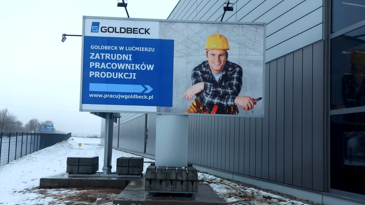 Billboard z reklamą firmy Goldbeck w zimowej scenerii, prezentujący pracownika w kasku i narzędziami, ogłaszający rekrutację pracowników produkcji.