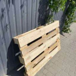 Drewniana paleta euro, ustawiona ukośnie na brukowanej powierzchni, z bluszczem pnącym się po metalowej ścianie w tle. Paleta o jasnym drewnie, widoczne oznaczenia EUR. Cień palety na bruku.