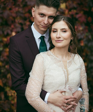 Uśmiechnięta para: panna młoda w sukni z długim rękawem ozdobioną cekinami i pan młody w bordowym garniturze z zielonym krawatem, pozują na tle jesiennych liści.