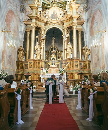 Para młoda siedzi tyłem do kamery podczas ceremonii ślubnej w bogato zdobionym kościele w Warszawie. Czerwony dywan prowadzi do ołtarza, a ławki są udekorowane białymi wstążkami i kwiatami.