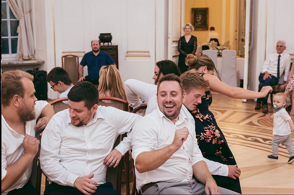 Rozbawieni goście w białych koszulach siedzą na krzesłach podczas uroczystości, w tle tańcząca para i małe dziecko z smoczkiem.