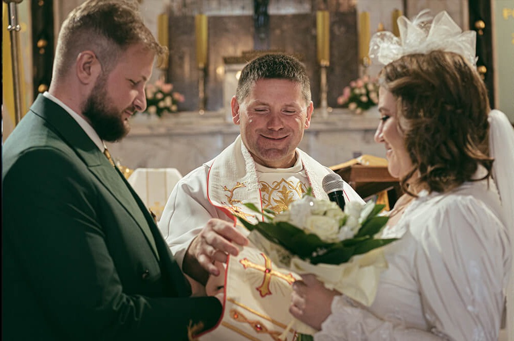 Panna młoda z bukietem i pan młody podczas błogosławieństwa przez księdza w kościele, widoczne zdobienia na ornacie i mikrofon.