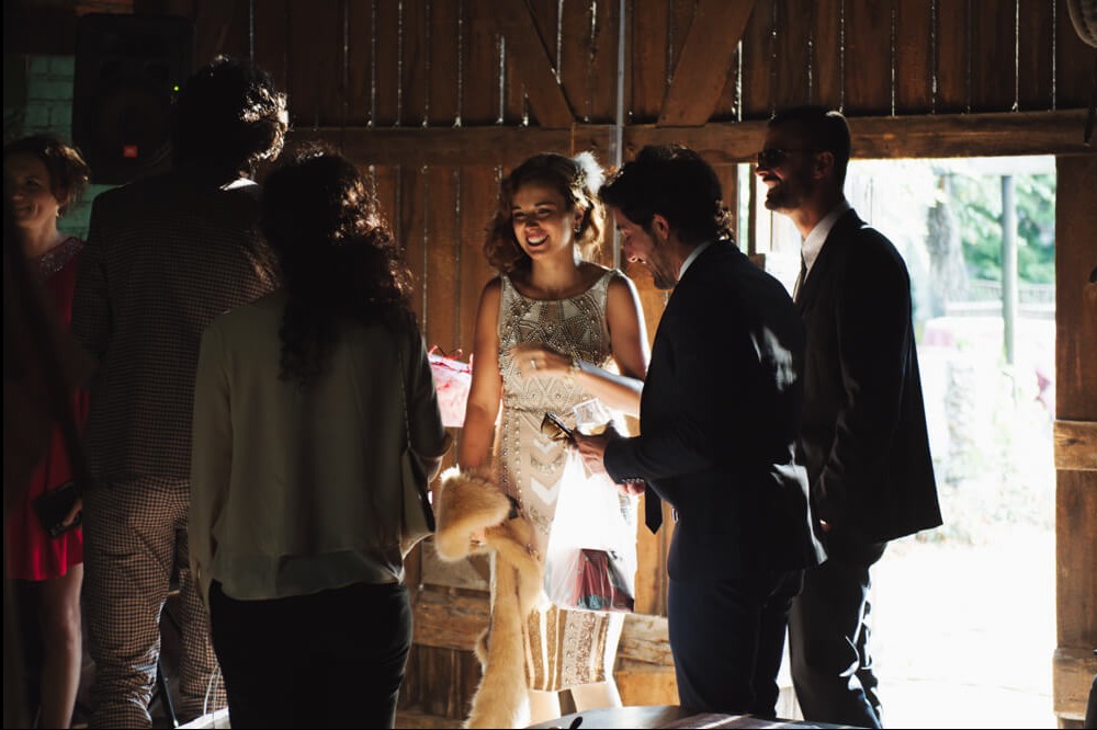 Panna młoda w sukni z aplikacjami, uśmiechnięta, otoczona gośćmi w rustykalnej stodole podczas przyjęcia weselnego.