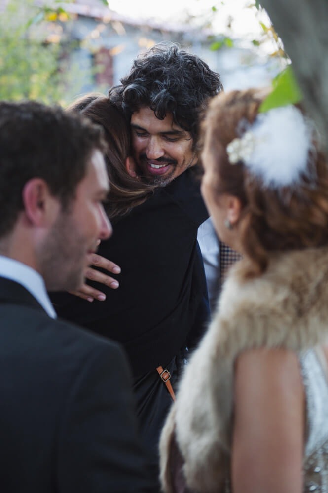 Uśmiechnięty mężczyzna z brodą w czarnej koszuli obejmuje kobietę podczas uroczystości, widoczna para w garniturach i kobieta w futrzanym szalu.