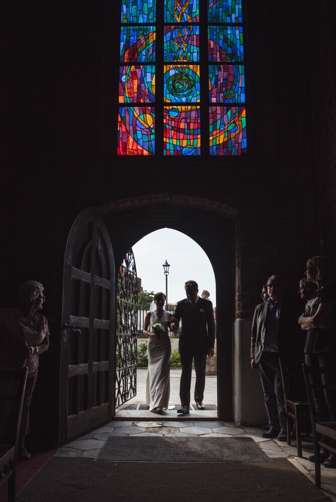 Panna młoda prowadzona przez ojca wchodzi do kościoła przez gotyckie drzwi, nad nimi kolorowy witraż, w tle widoczna latarnia.