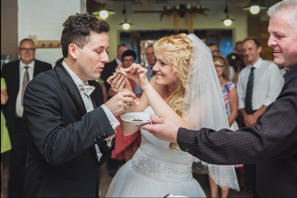 Para młoda w trakcie wesela, karmiąca się tortem, uśmiechnięta panna młoda z blond lokami i welonem, elegancki pan młody w smokingu, w tle goście weselni.