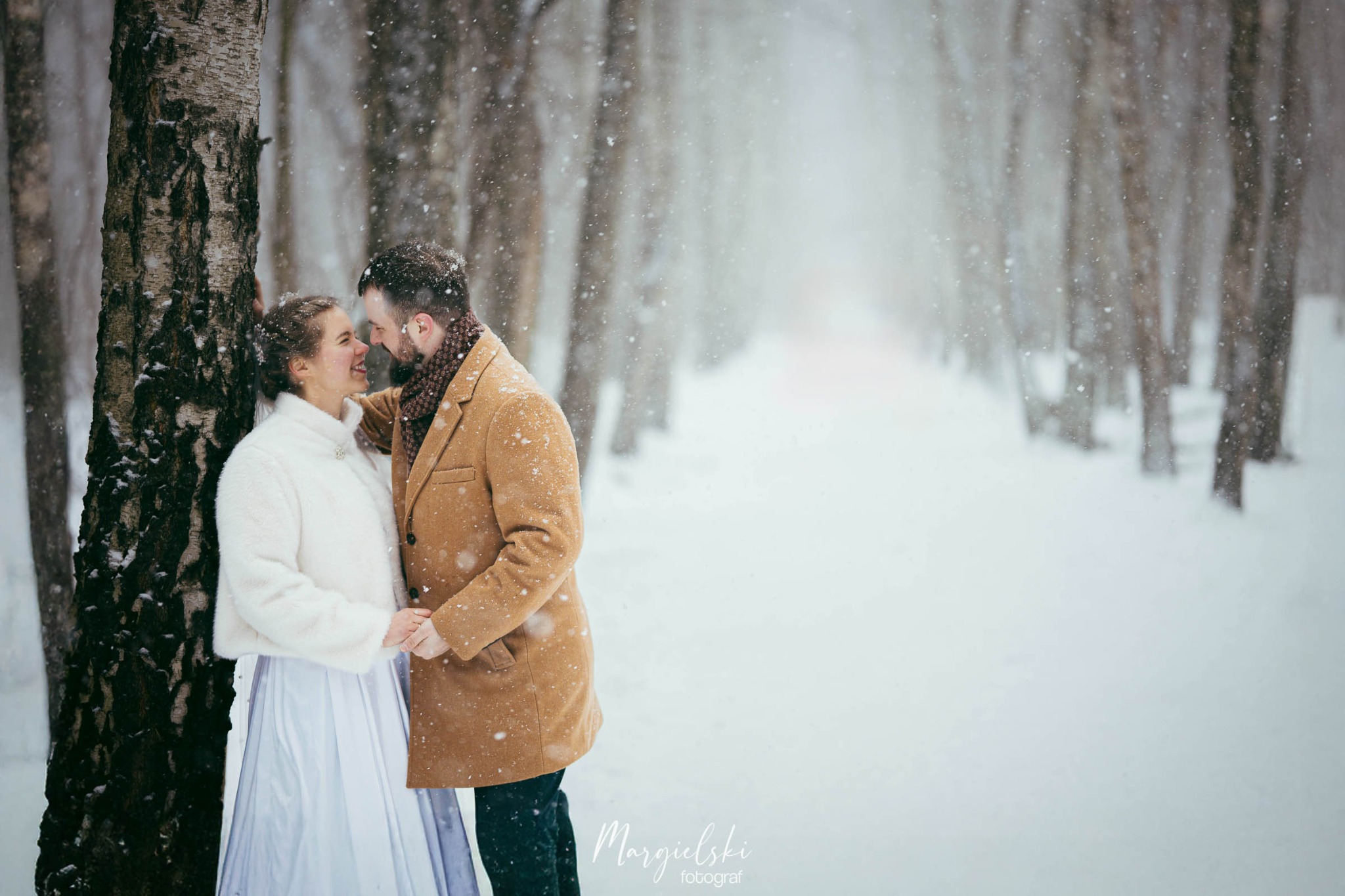 Zimowa sesja narzeczeńska: para w objęciach pod drzewem, padający śnieg tworzy romantyczną atmosferę. Ona w białym futerku, on w brązowym płaszczu.