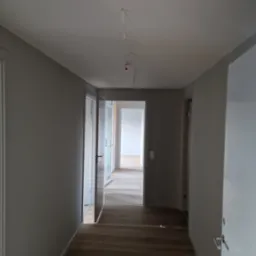 Wnętrze korytarza po remoncie z szarymi ścianami, drewnianą podłogą i otwartymi drzwiami, widać wystające kable z sufitu.