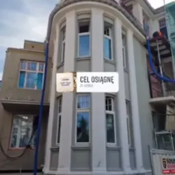 Renowacja elewacji budynku z charakterystyczną wieżyczką, widoczne rusztowania i pracownik z niebieskim wężem, szaro-biała kolorystyka budynku.