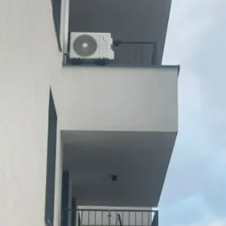 Zewnętrzna jednostka klimatyzacji zamontowana na balkonie nowoczesnego budynku mieszkalnego o minimalistycznej architekturze, widok z dołu na tle pochmurnego nieba.