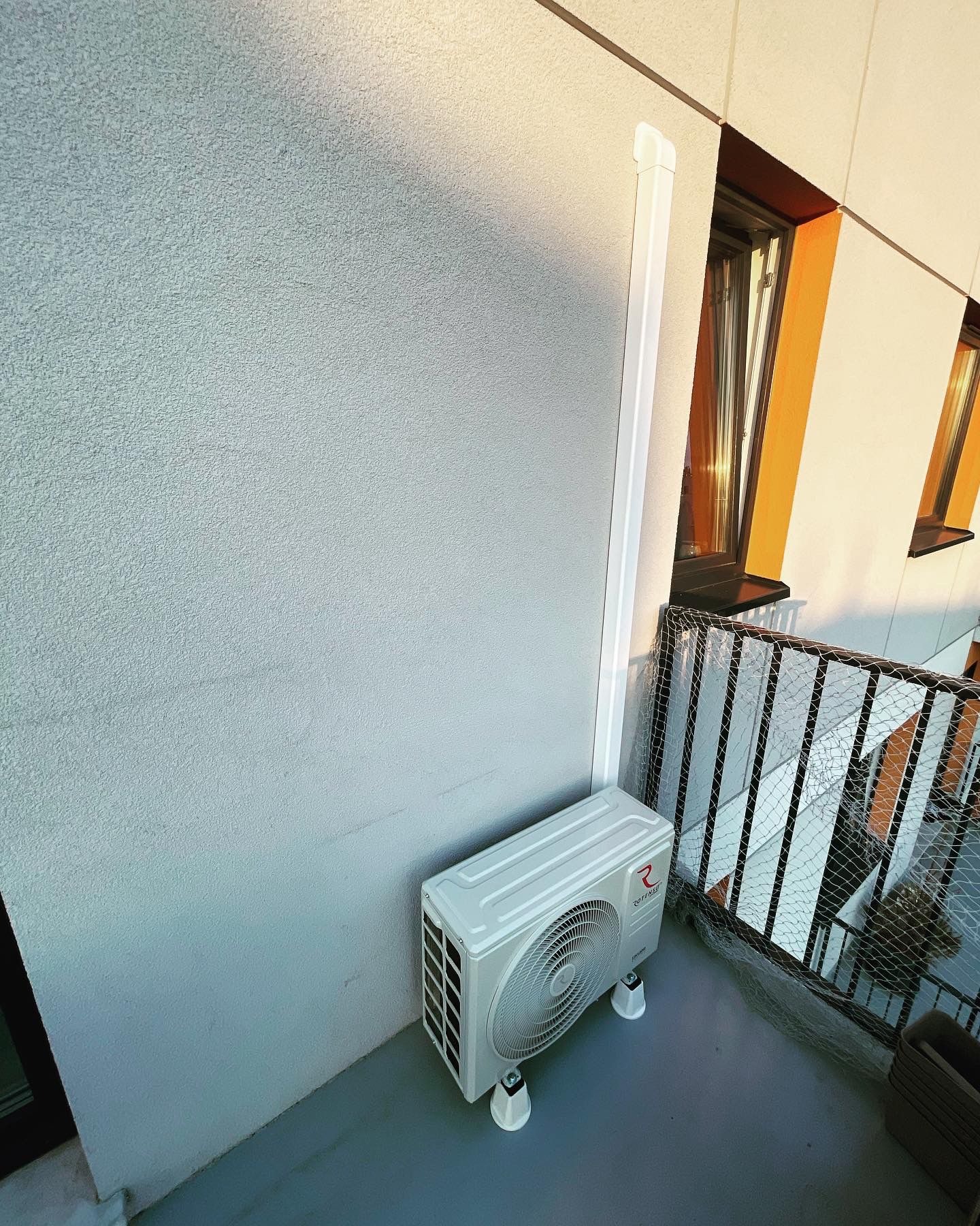 Zewnętrzna jednostka klimatyzacyjna zamontowana na balkonie z widoczną rurą odprowadzającą, zabezpieczona siatką balkonowa, oświetlona popołudniowym słońcem.