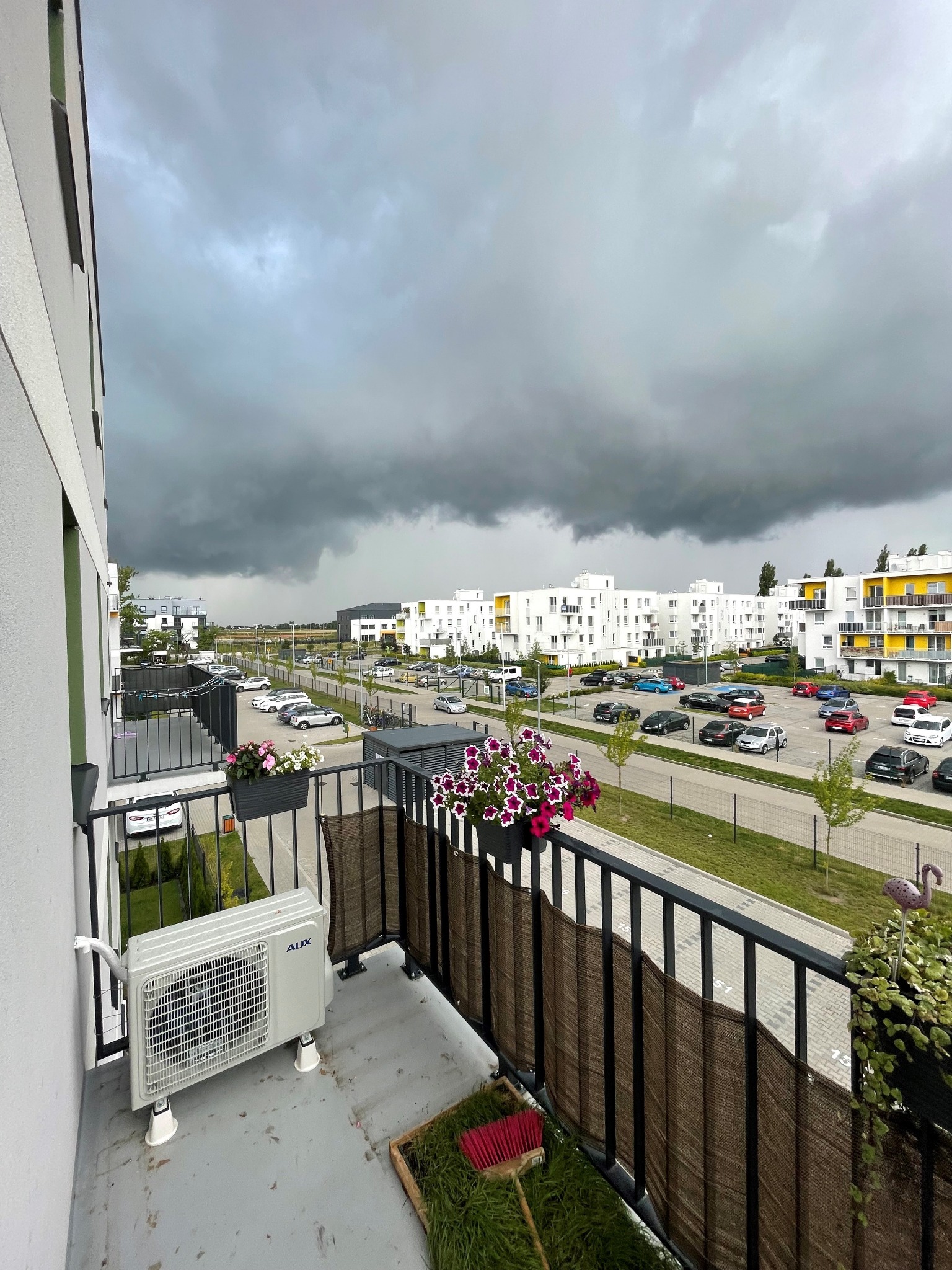 Klimatyzator AUX zamontowany na balkonie nowego bloku mieszkalnego, widok na parking i zbierające się ciemne chmury.