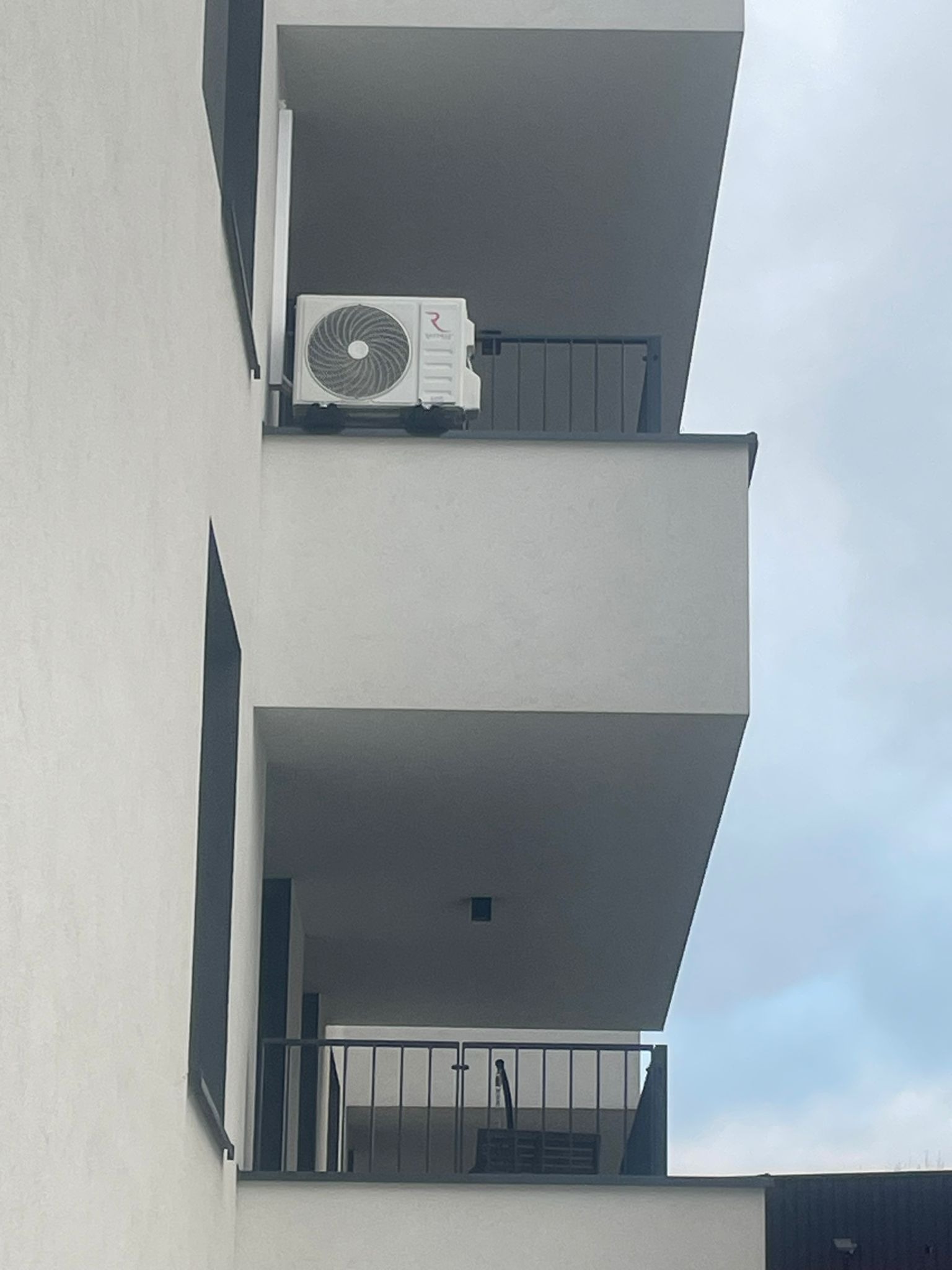 Zewnętrzna jednostka klimatyzacji zamontowana na balkonie nowoczesnego budynku mieszkalnego o minimalistycznej architekturze, widok z dołu na tle pochmurnego nieba.