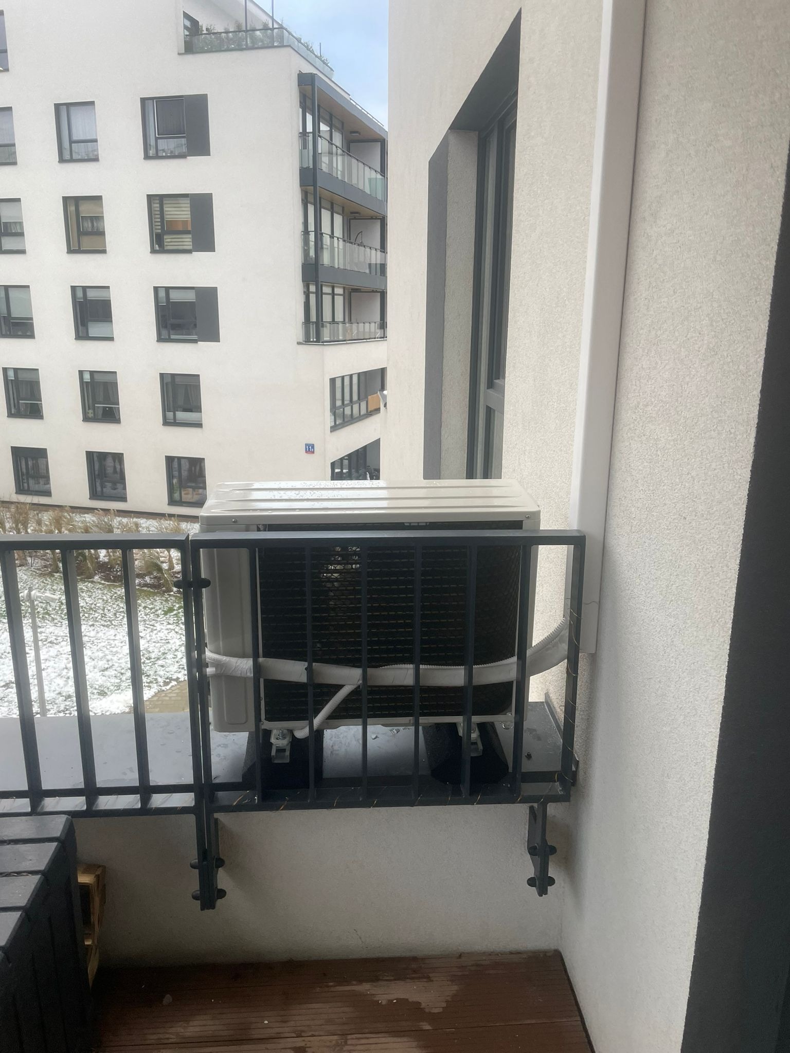Jednostka zewnętrzna klimatyzacji zamontowana na balkonie w bloku mieszkalnym, widoczne przewody i uchwyty montażowe, w tle ośnieżony krajobraz miejski.