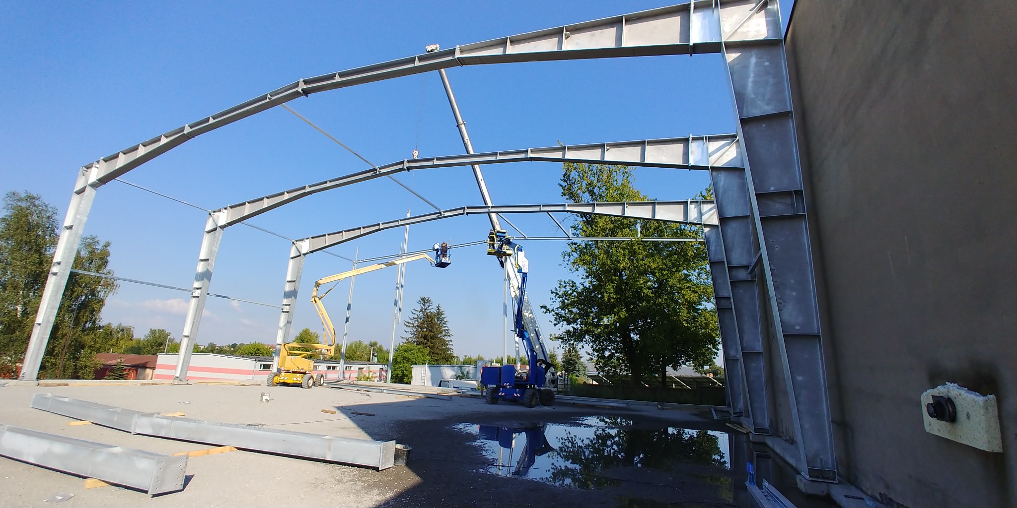 Montaż stalowej konstrukcji hali: ekipa na podnośnikach koszowych łączy elementy szkieletu, widoczne niepołączone profile leżące na placu budowy, odbicie konstrukcji w kałuży.
