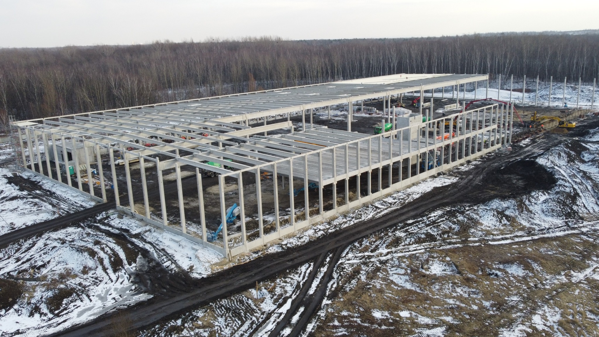 Budowa hali logistycznej w zimowej scenerii, widok z lotu ptaka na szkielet konstrukcji, częściowo pokryty śniegiem teren budowy, w tle las.