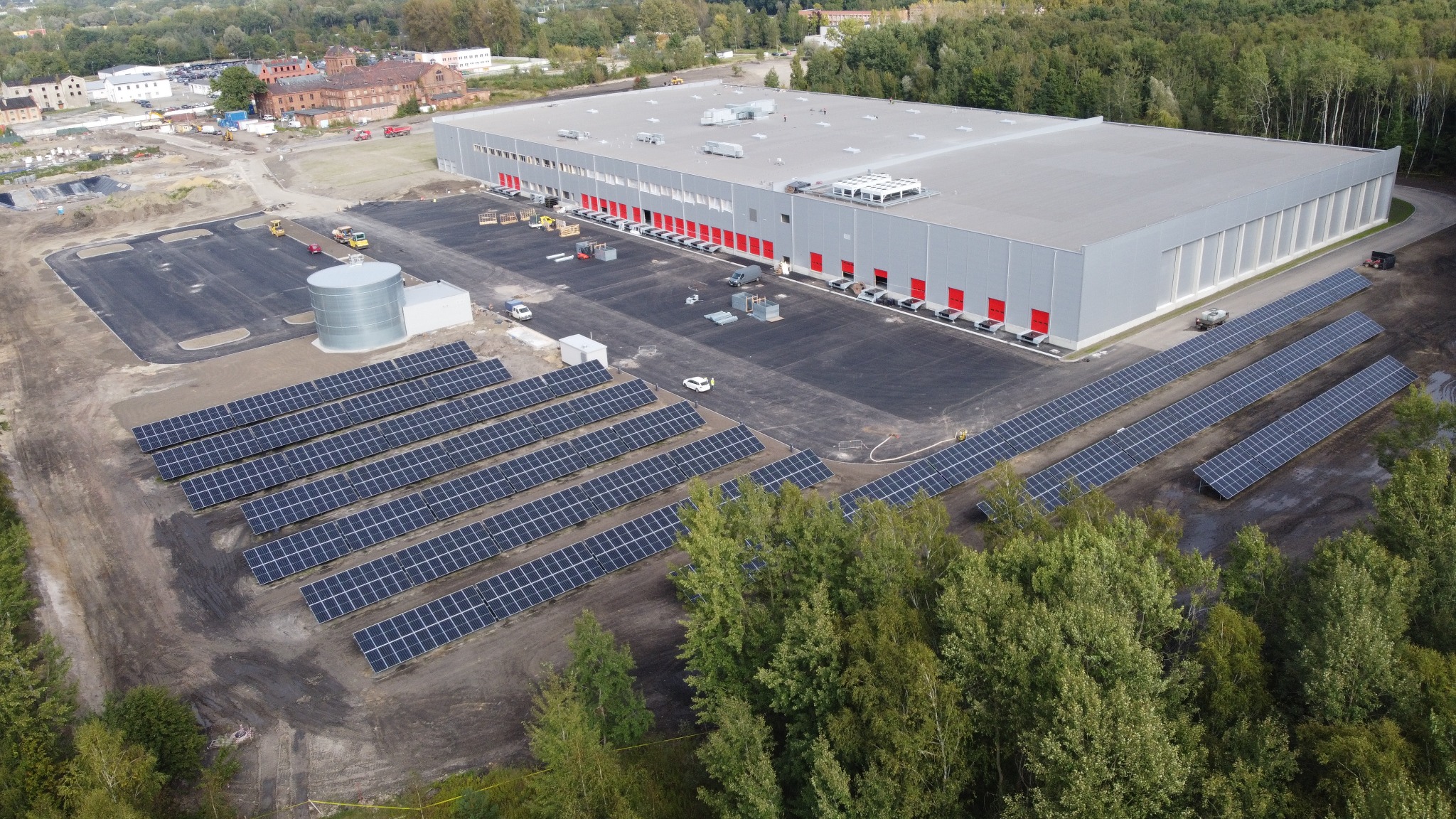 Szeroki plan z lotu ptaka na ukończone Centrum Logistyczne Transgourmet o powierzchni 40000m2, z panelami słonecznymi na pierwszym planie i budynkiem magazynowym w tle, widoczne czerwone bramy dokowe