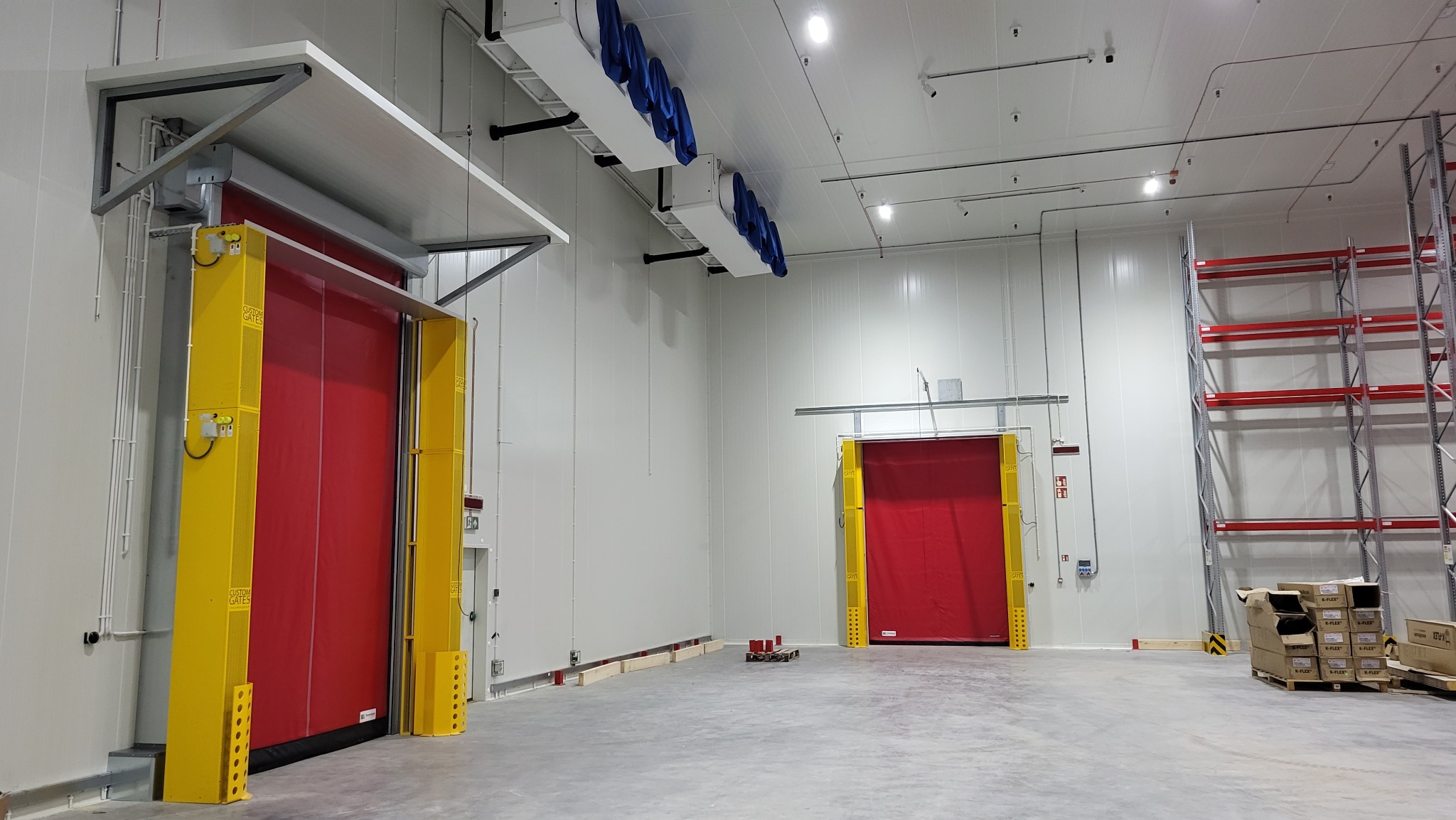 Wnętrze hali magazynowej z dwoma czerwonymi, szybkobieżnymi bramami przemysłowymi z żółtymi prowadnicami, widoczne regały magazynowe i system wentylacji sufitowej.