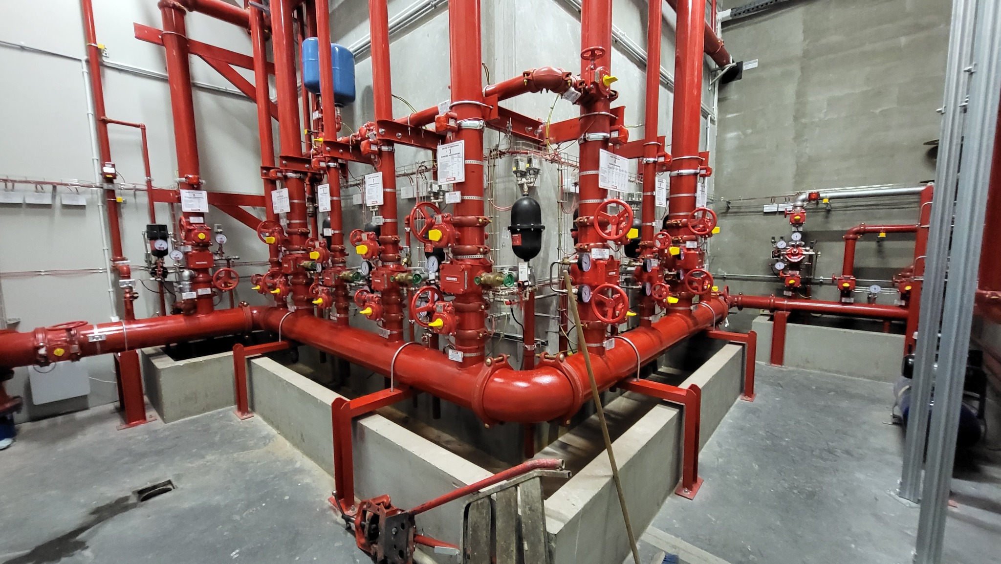 Kompleksowa instalacja przeciwpożarowa z czerwonymi rurami, zaworami i manometrami w nowym budynku, widoczne podpory i fragment drabiny.