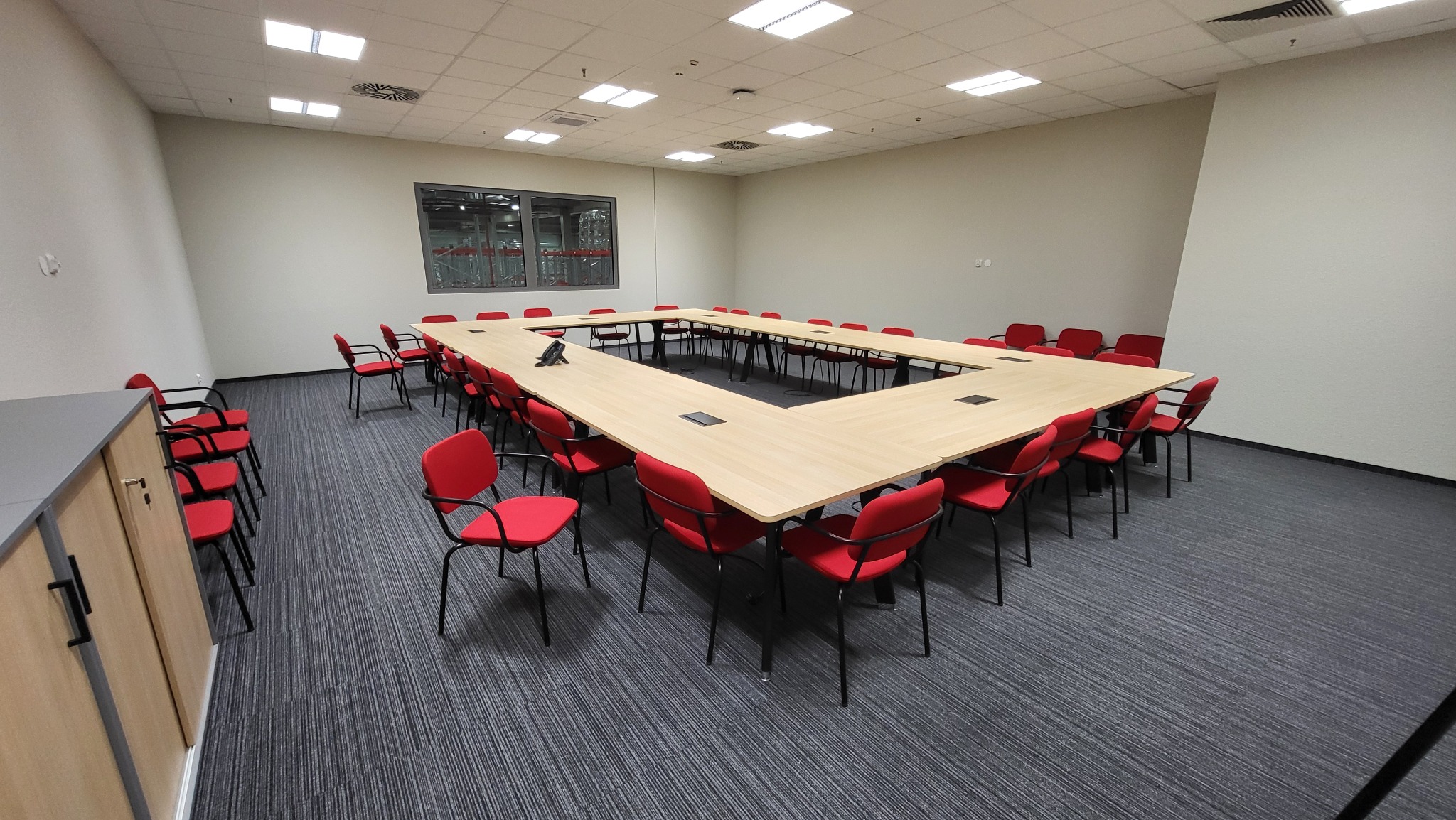 Sala konferencyjna z dużym stołem w kształcie litery U i czerwonymi krzesłami, widok na okno z elementami konstrukcji hali w tle.